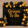 Groom to be party decoration with black and gold balloons, black foil curtain backdrop, golden Groom To Be foil banner, gold confetti balloons and a bottle foil balloon.