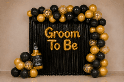 Groom To Be Black Gold Decoration with Bottle Foil Balloon Groom to be party decoration with black and gold balloons, black foil curtain backdrop, golden Groom To Be foil banner, gold confetti balloons and a bottle foil balloon.