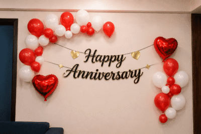 Happy Anniversary decoration with red and white balloon clusters, black and gold cursive anniversary banner, and red heart foil balloons on a home wall backdrop.